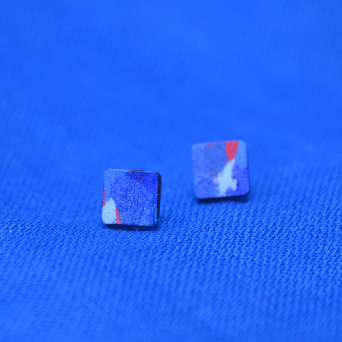 A pair of wooden square stud earrings with a painted blue and red pattern on a textured blue background.