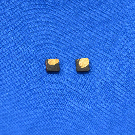A pair of square stud earrings with a black wood-grain finish and a textured gold painted accent, displayed on a vibrant blue textured fabric background.