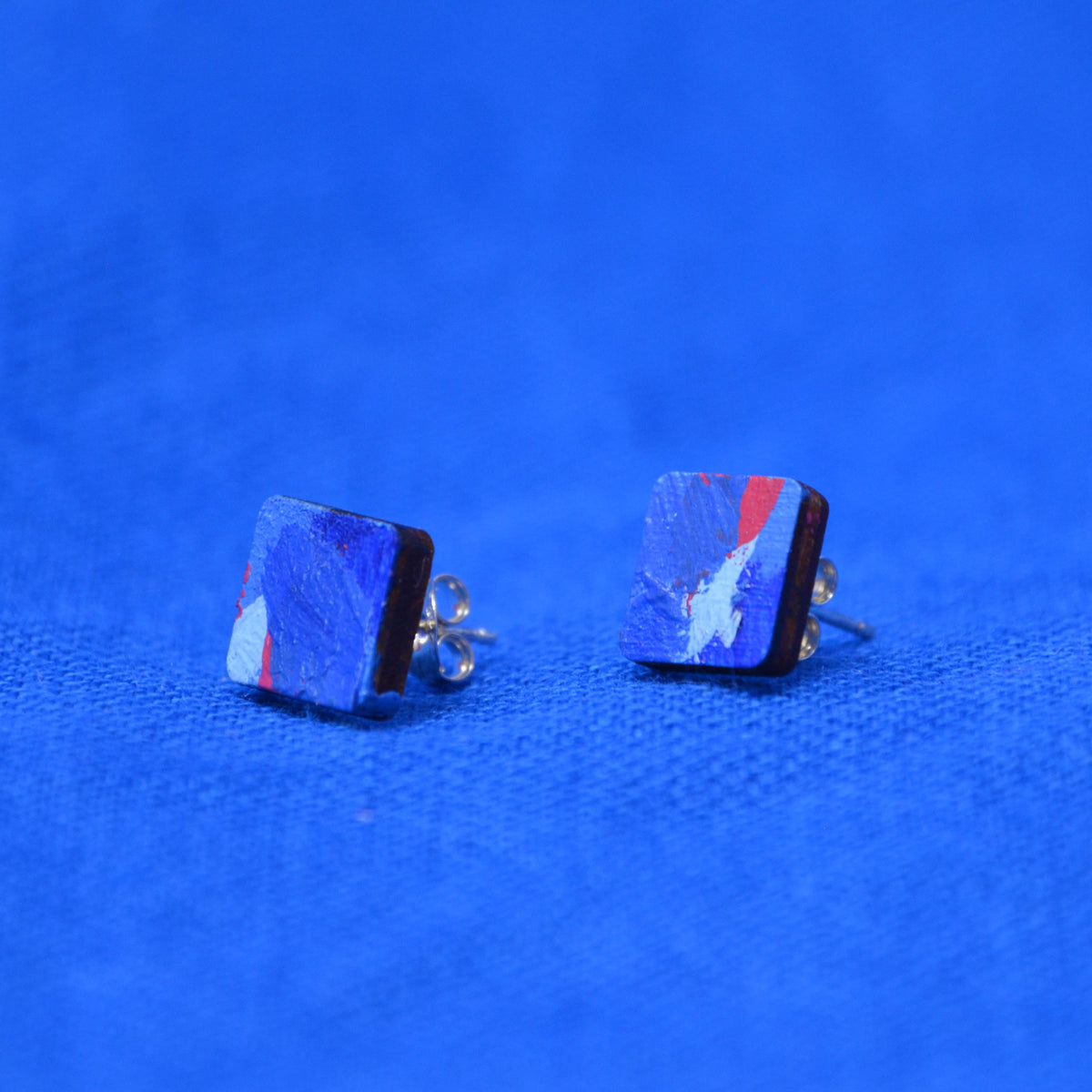 A pair of wooden square stud earrings with a painted blue and red pattern on a textured blue background.