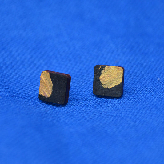 A pair of square stud earrings with a black wood-grain finish and a textured gold painted accent, displayed on a vibrant blue textured fabric background.