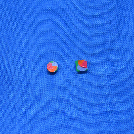 Two small multicolored square and round wooden earrings, displayed on a textured blue background.