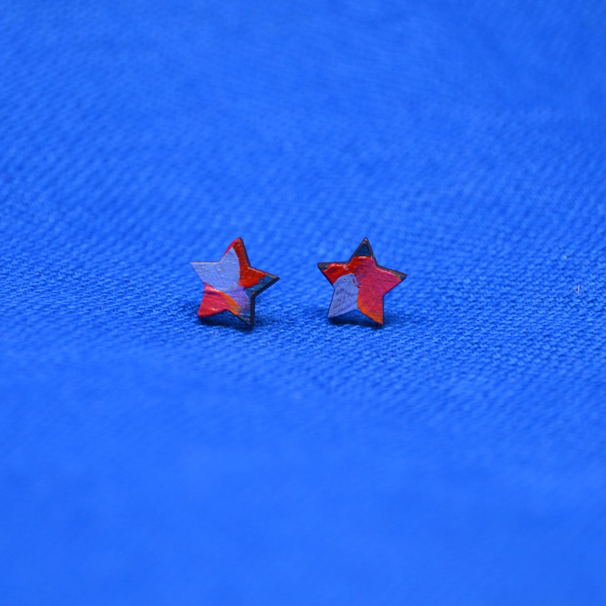 Two small wooden star-shaped stud earrings with a black, orange, red, pink and purple painted pattern, displayed on a textured blue background.