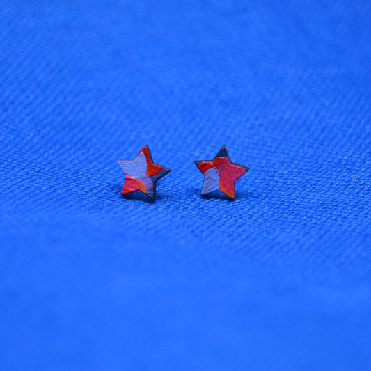 Two small wooden star-shaped stud earrings with a black, orange, red, pink and purple painted pattern, displayed on a textured blue background.