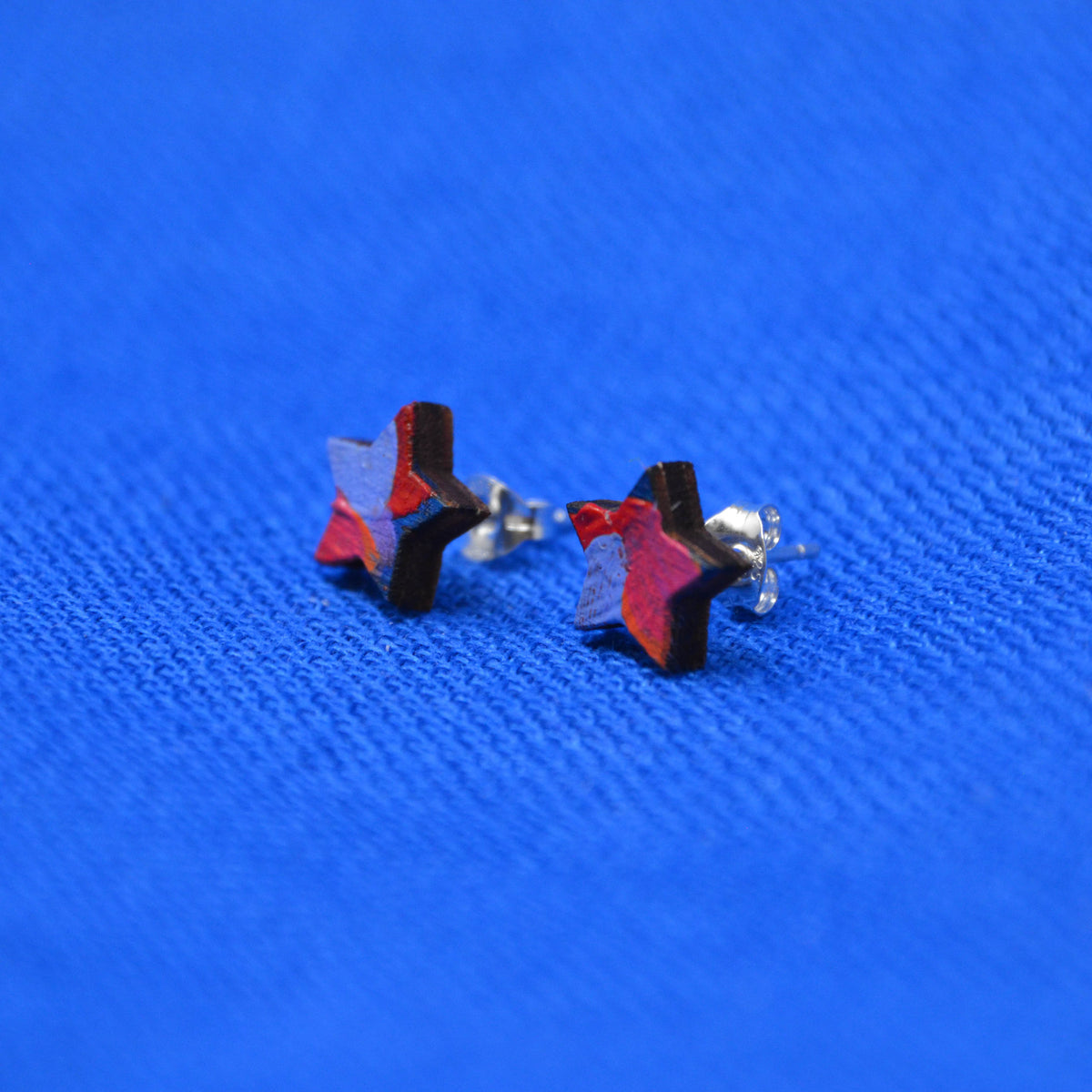 Two small wooden star-shaped stud earrings with a black, orange, red, pink and purple painted pattern, displayed on a textured blue background.