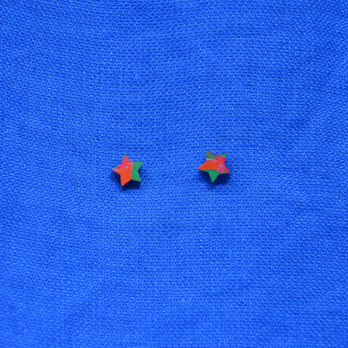 A pair of small star-shaped stud earrings with a red-orange and green color-blocked design, displayed on a textured blue fabric background.