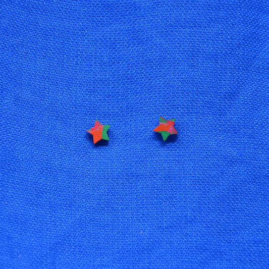 A pair of small star-shaped stud earrings with a red-orange and green color-blocked design, displayed on a textured blue fabric background.