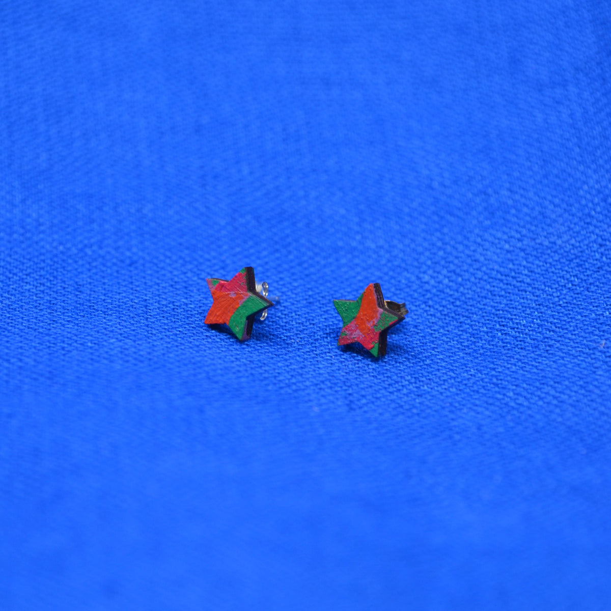 Two small star-shaped stud earrings with a red, orange and green painted pattern, displayed on a textured blue background.