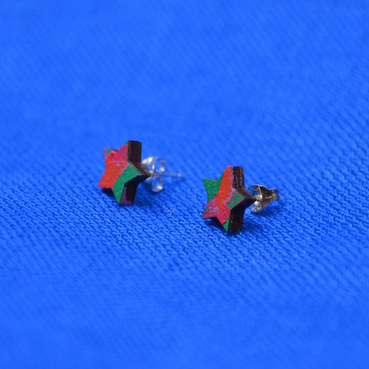A pair of small star-shaped stud earrings with red, green, and dark brown wooden sides and sterling silver posts, placed on a textured bright blue surface.