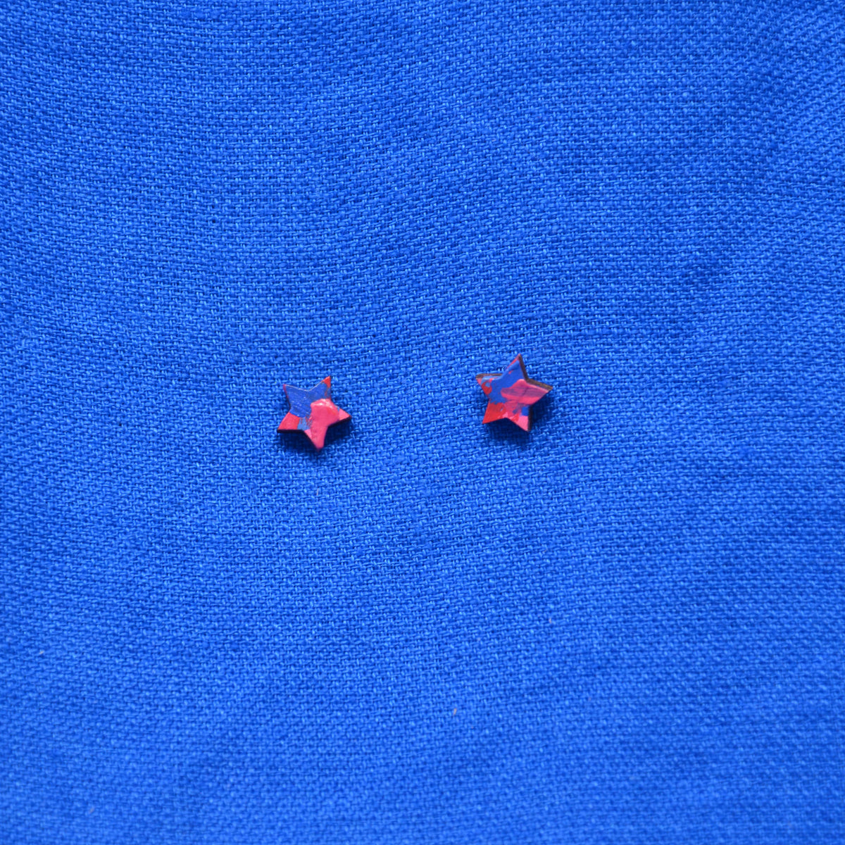 Two small wooden star-shaped stud earrings with a pink and blue painted pattern, displayed on a textured blue background.