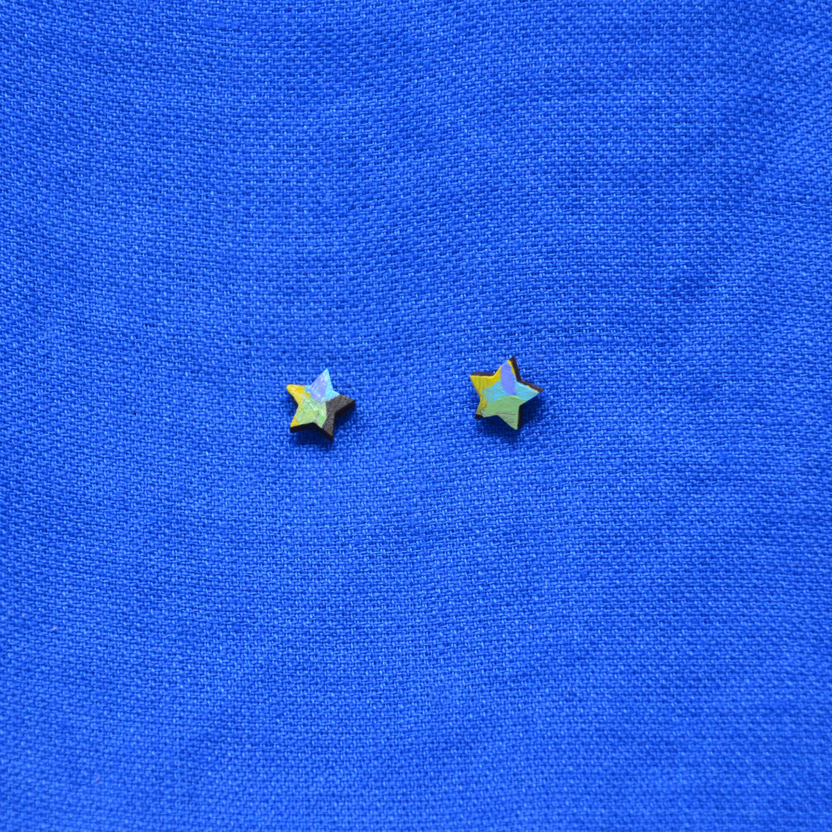 Two small star-shaped stud earrings with a black, yellow, green, purple and blue painted pattern, displayed on a textured blue background.