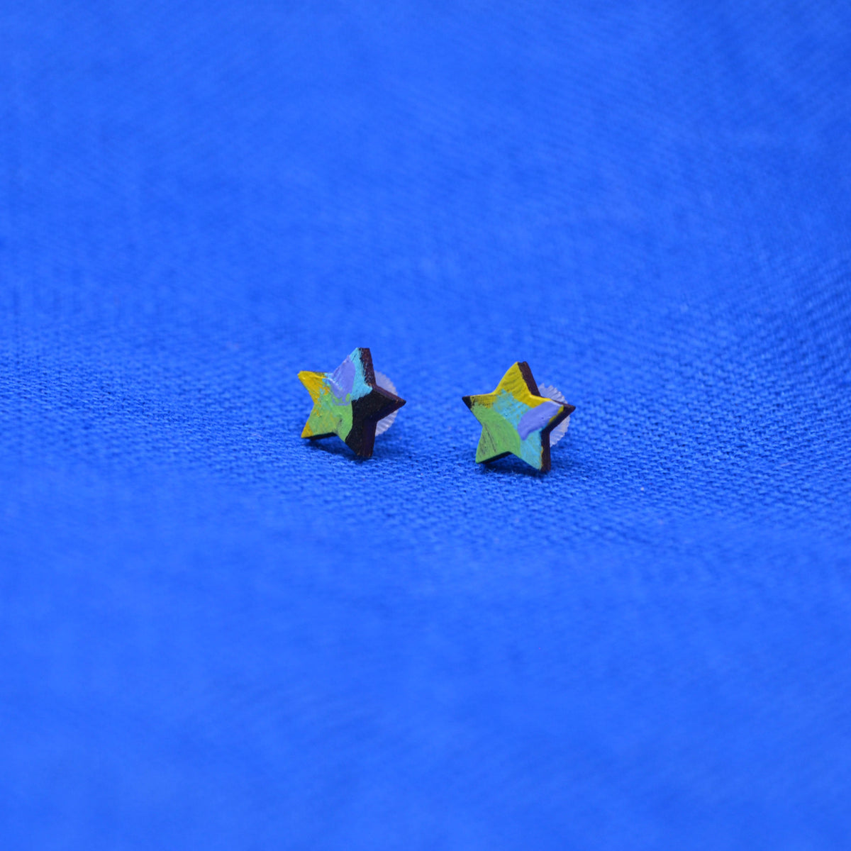 Two small star-shaped stud earrings with a black, yellow, green, purple and blue painted pattern, displayed on a textured blue background.