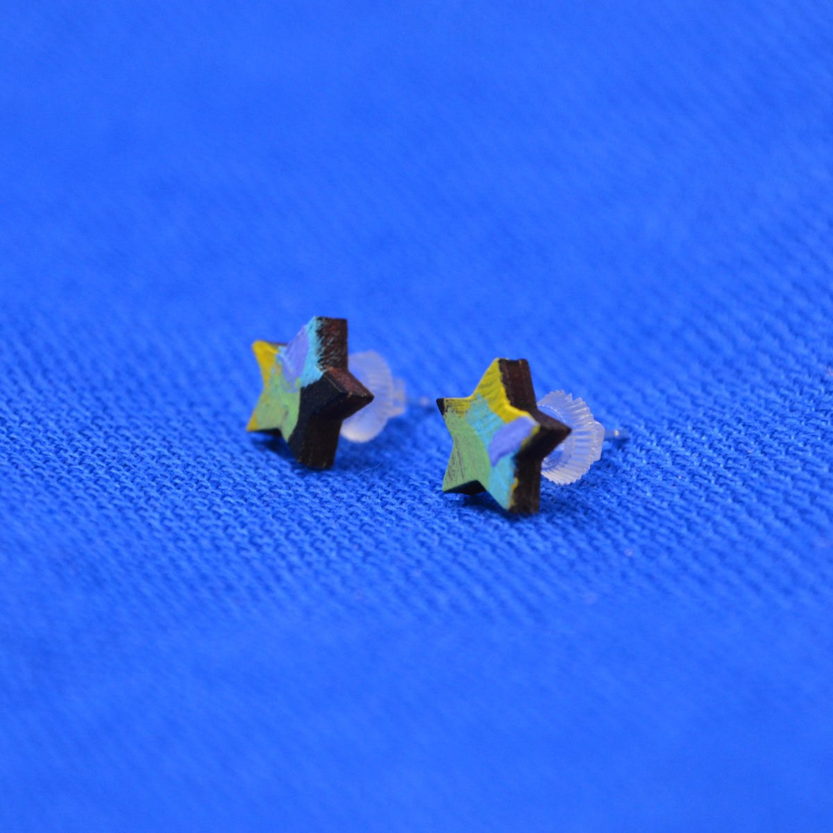 Two small star-shaped stud earrings with a black, yellow, green, purple and blue painted pattern, displayed on a textured blue background.