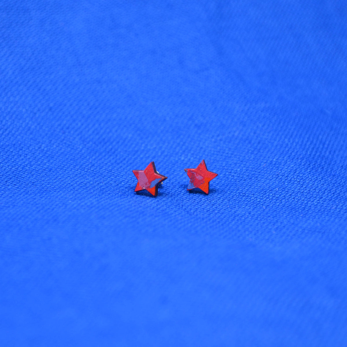 A pair of small star-shaped stud earrings in a vibrant red-orange color, displayed on a textured blue fabric background.