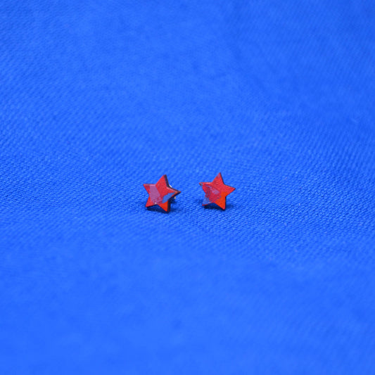 A pair of small star-shaped stud earrings in a vibrant red-orange color, displayed on a textured blue fabric background.