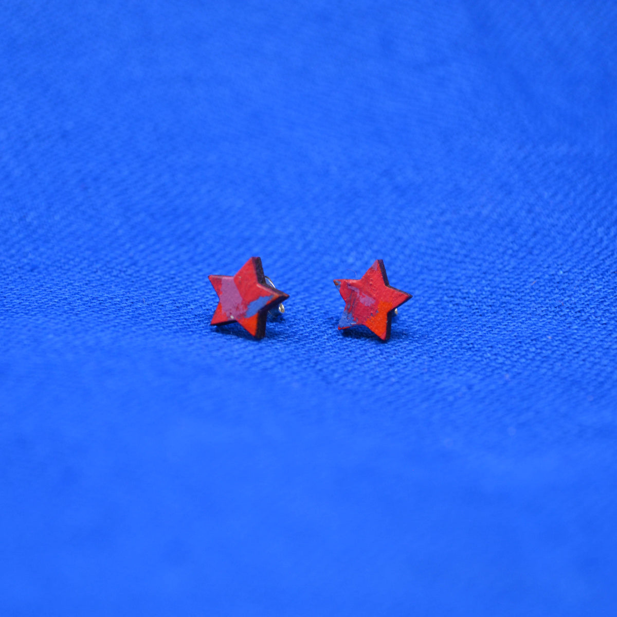 A pair of small red star-shaped stud earrings on a textured bright blue background.