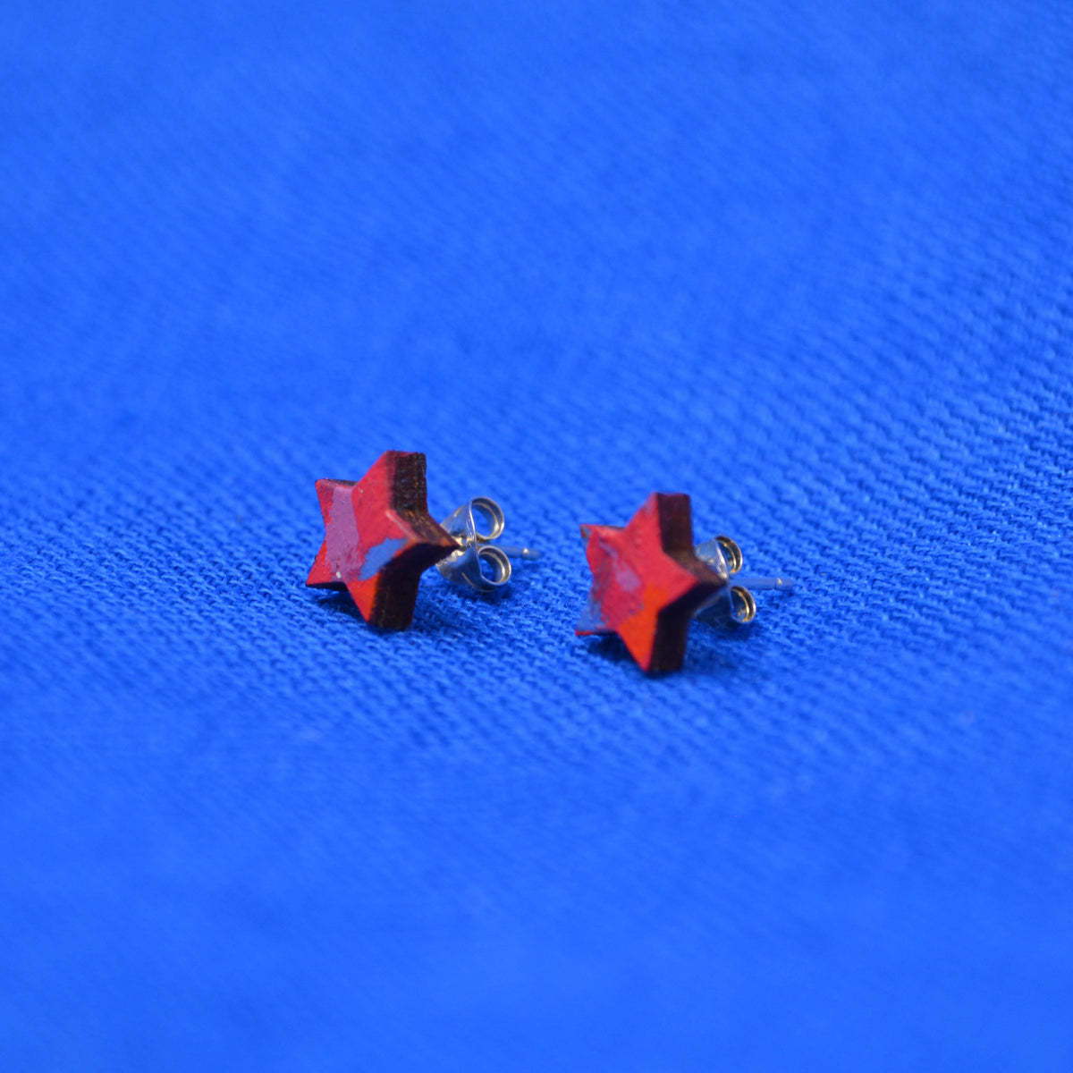 A pair of red star-shaped stud earrings with sterling silver posts, displayed on a textured blue background.