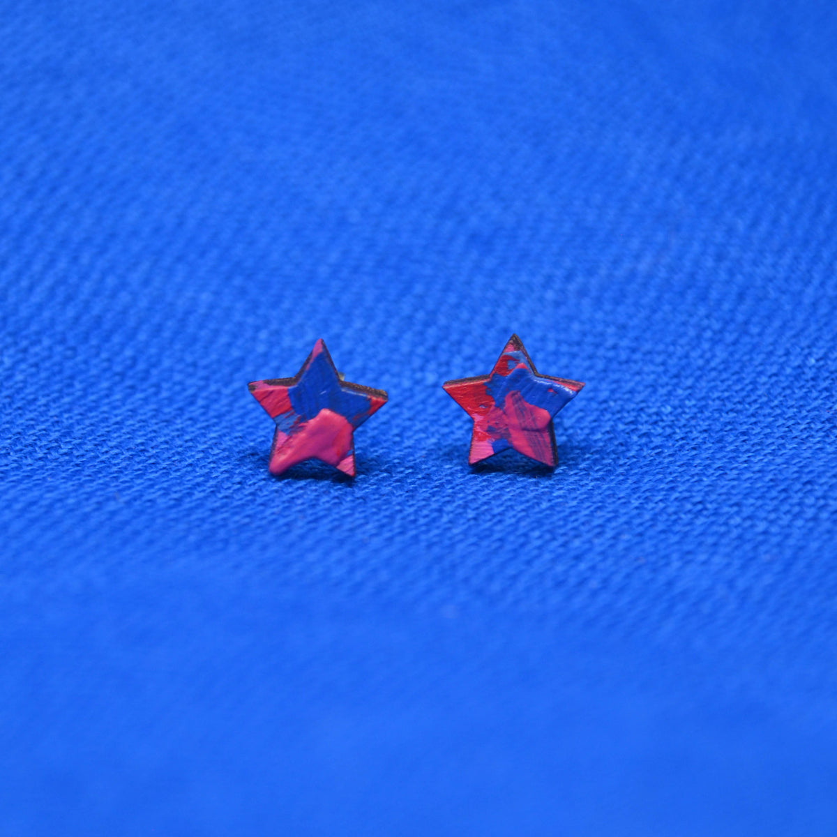 Two small star-shaped stud earrings with a pink and blue painted pattern, displayed on a textured blue background.