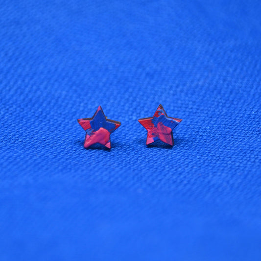 Two small star-shaped stud earrings with a pink and blue painted pattern, displayed on a textured blue background.