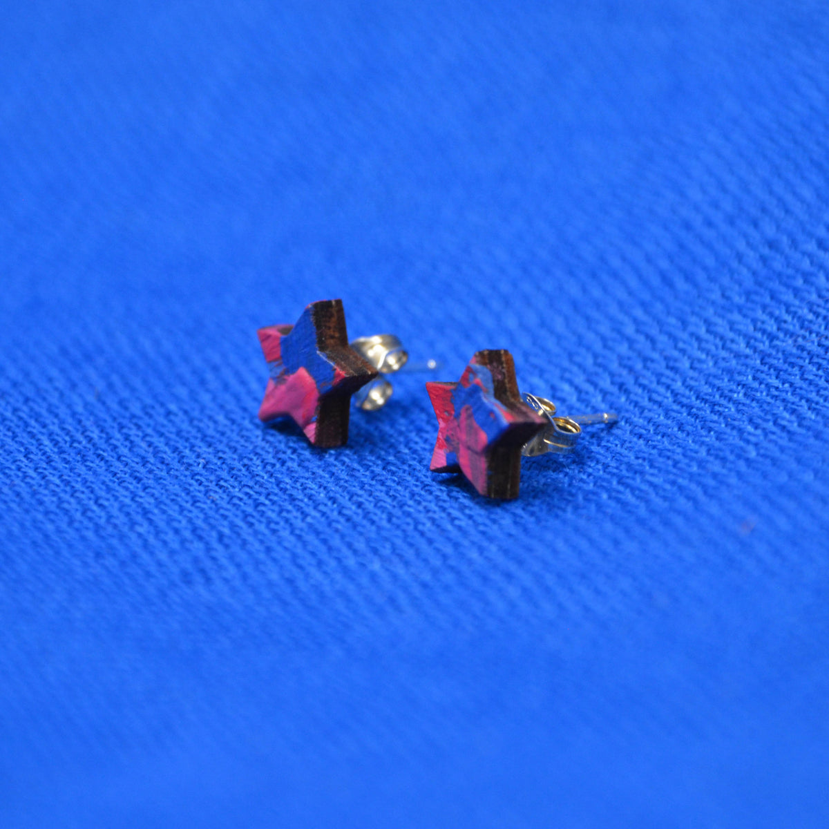 A pair of star-shaped stud earrings with a marbled pink, red, on a wooden base, shown on a textured blue background.