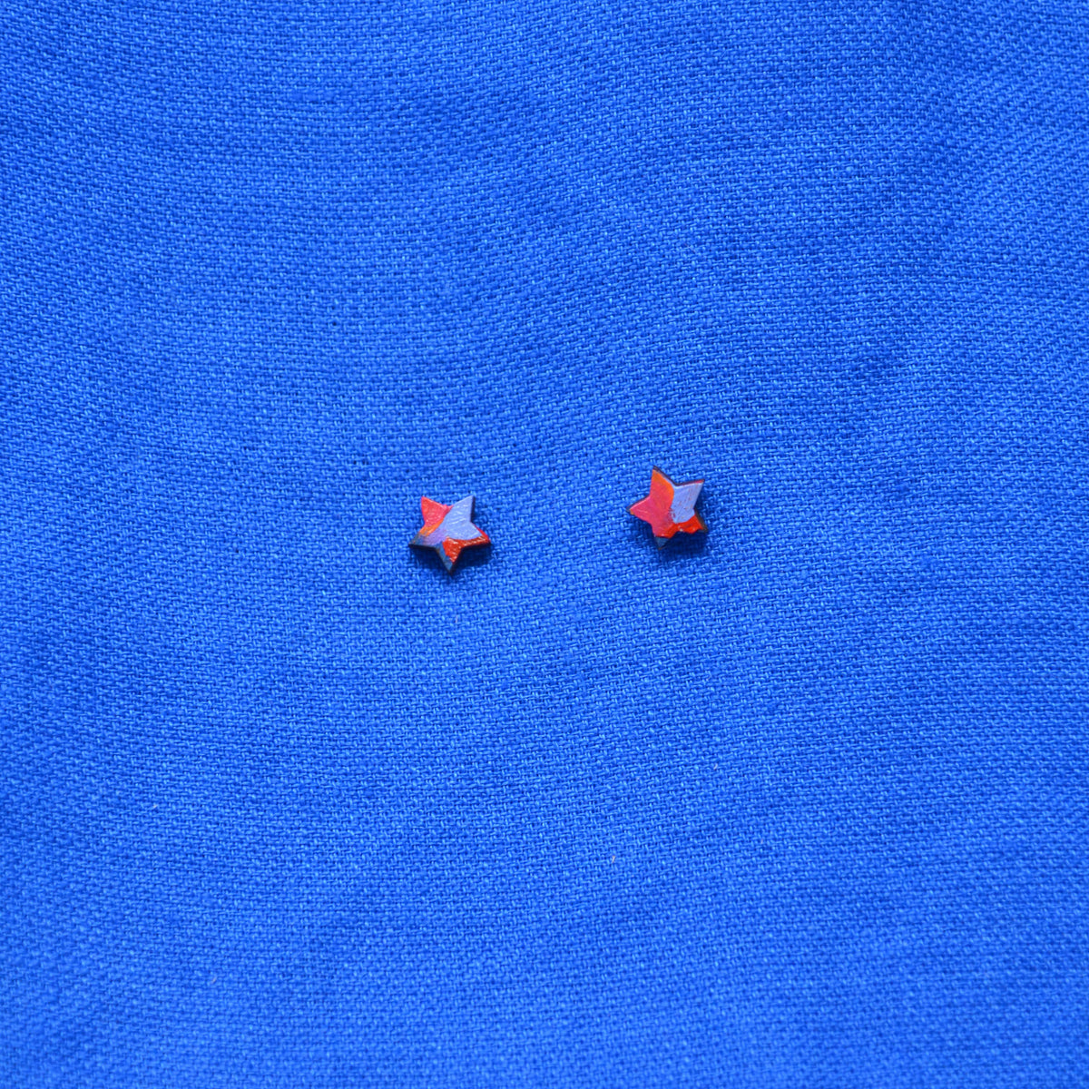 Two small wooden star-shaped stud earrings with a black, orange, red, pink and purple painted pattern, displayed on a textured blue background.