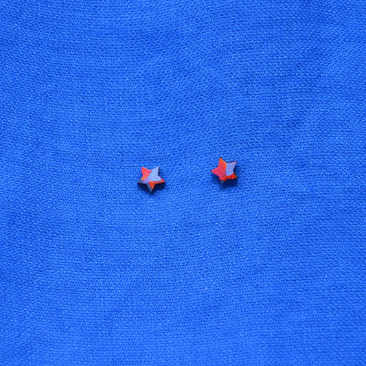 Two small wooden star-shaped stud earrings with a black, orange, red, pink and purple painted pattern, displayed on a textured blue background.