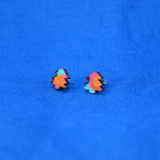 A pair of small multicolor stud earrings shaped like stylized trees, featuring orange, pink, and light blue with black outlines, placed on a textured blue fabric background.