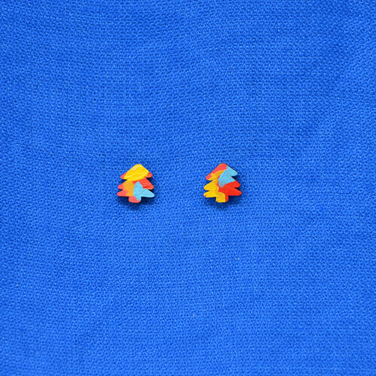 A pair of small multicolor tree stud earrings with yellow, orange, red, and light blue painted detail, displayed on a textured blue fabric background.
