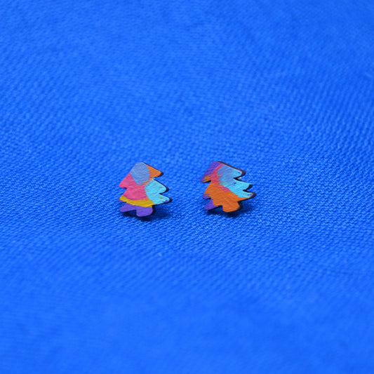 A pair of small, colorful tree-shaped stud earrings with a painted finish, displayed on a textured blue fabric background.