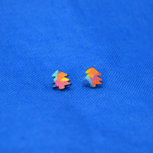 A pair of small, colorful Christmas tree stud earrings with a vibrant multicolor painted pattern, displayed on a textured blue fabric background.