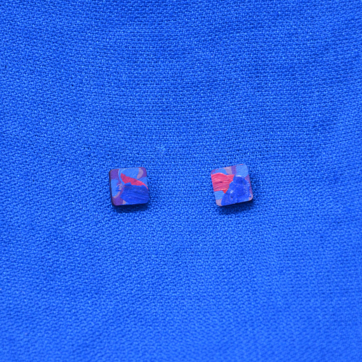 A pair of small square stud earrings with a marbled blue, purple, and red pattern, displayed on a textured blue fabric background.