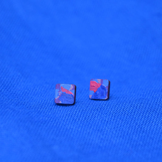 A pair of wooden square stud earrings with a painted blue, purple, and red pattern on a textured blue background.