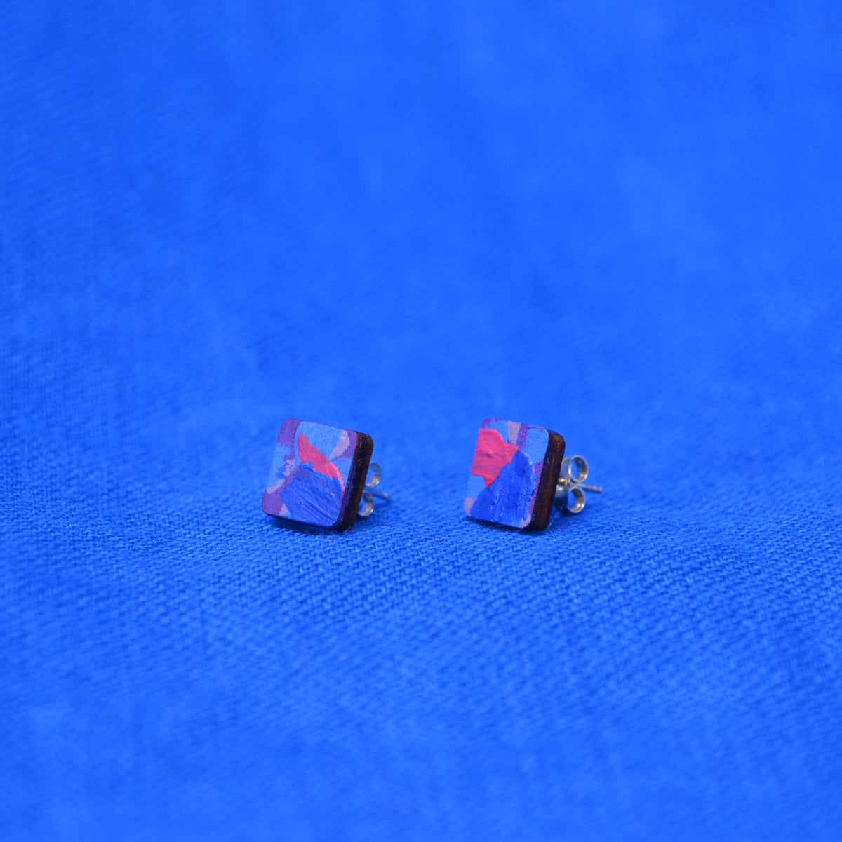 A pair of square stud earrings with abstract blue, purple, and red marbled patterns, displayed on a textured blue fabric background.