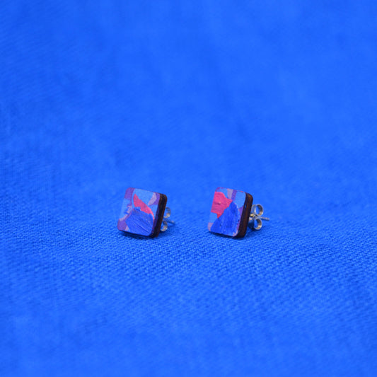 A pair of square stud earrings with abstract blue, purple, and red marbled patterns, displayed on a textured blue fabric background.