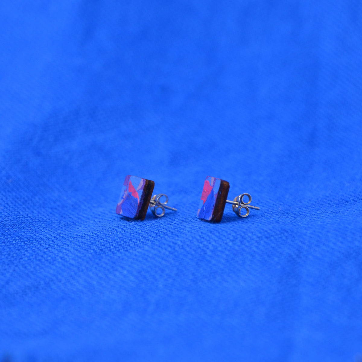 A pair of wooden square stud earrings with a painted blue, purple, and red pattern on a textured blue background.