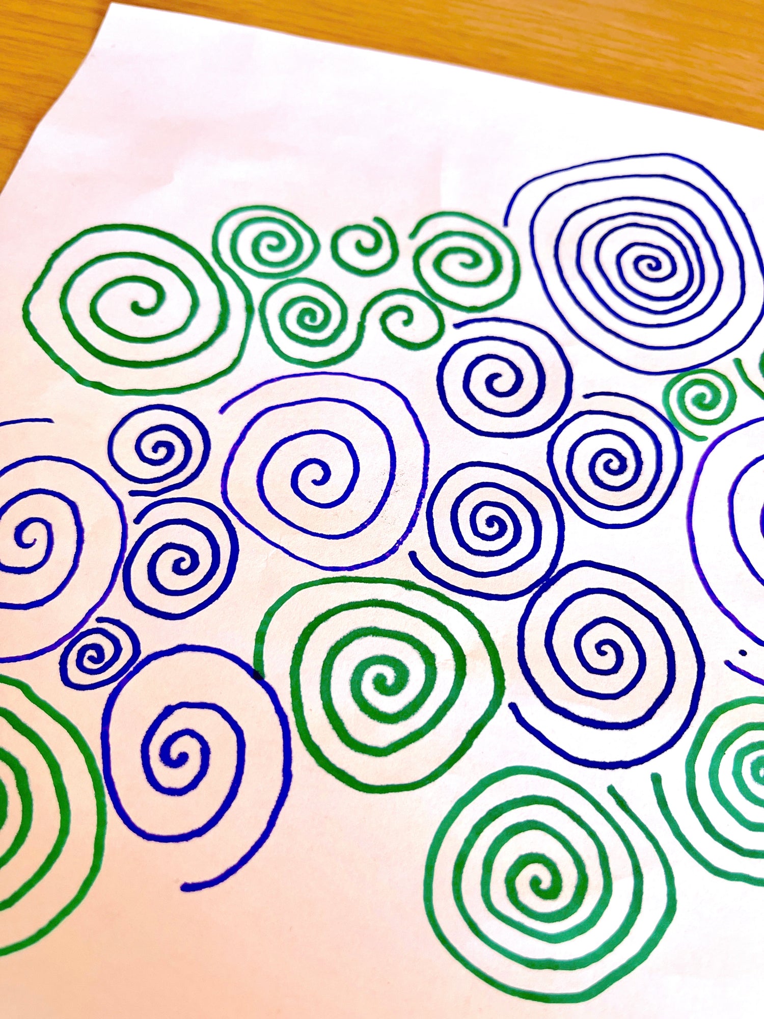 A close-up of a white sheet of paper covered with overlapping hand-drawn spiral patterns in green and blue, placed on a light wood surface.