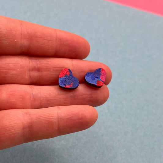 Two small heart-shaped earrings held between fingers against a multicolored background