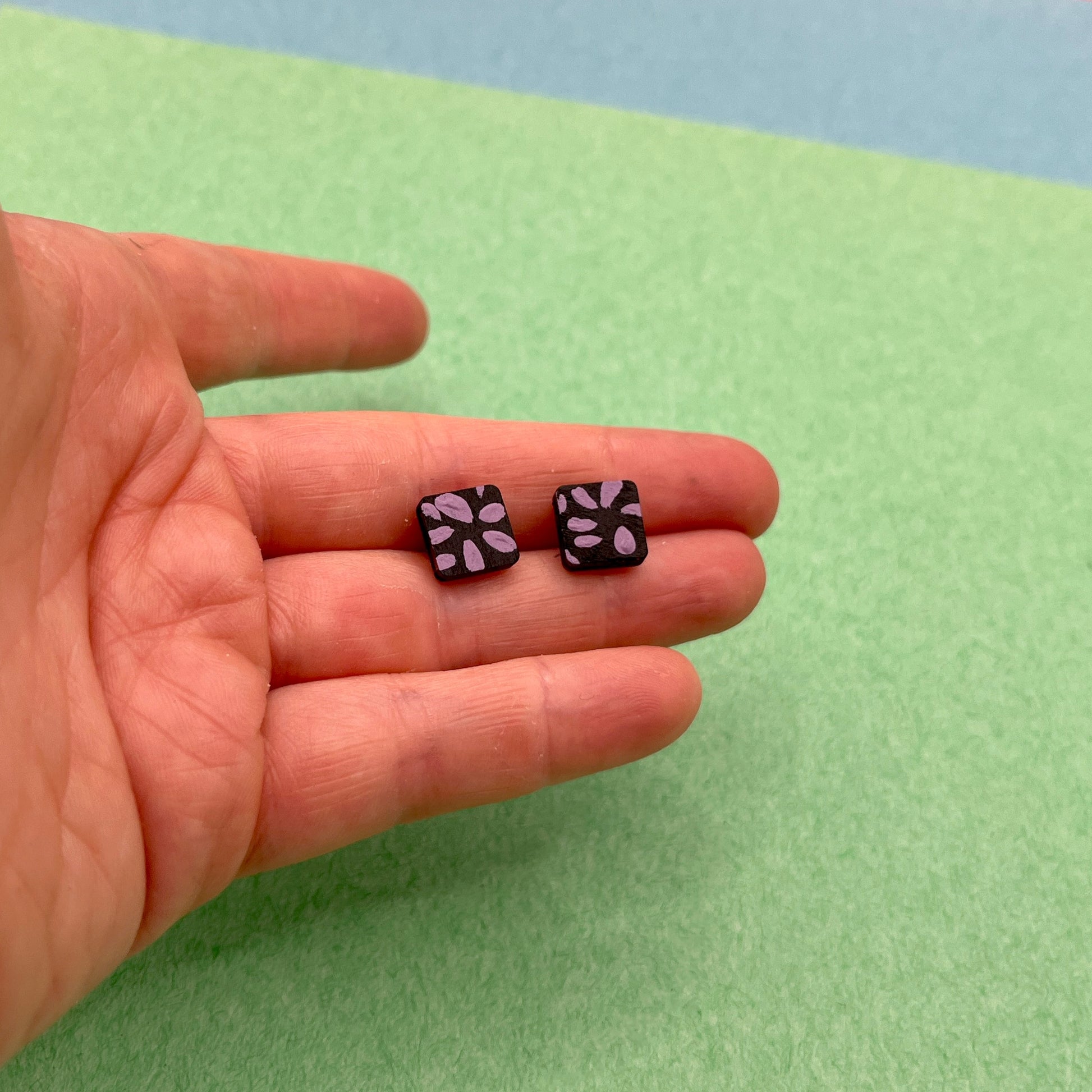 Two small black earrings with purple patterns held in a hand against a green background