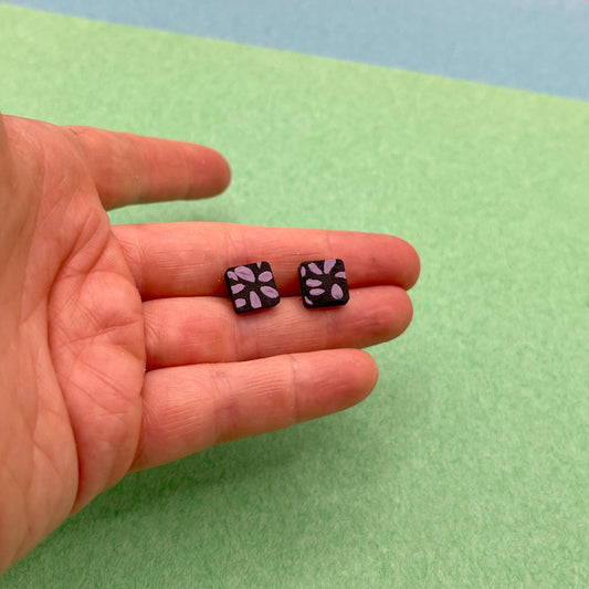 Two small black earrings with purple patterns held in a hand against a green background