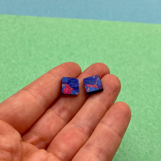Small square earrings held between fingers against a green and blue background